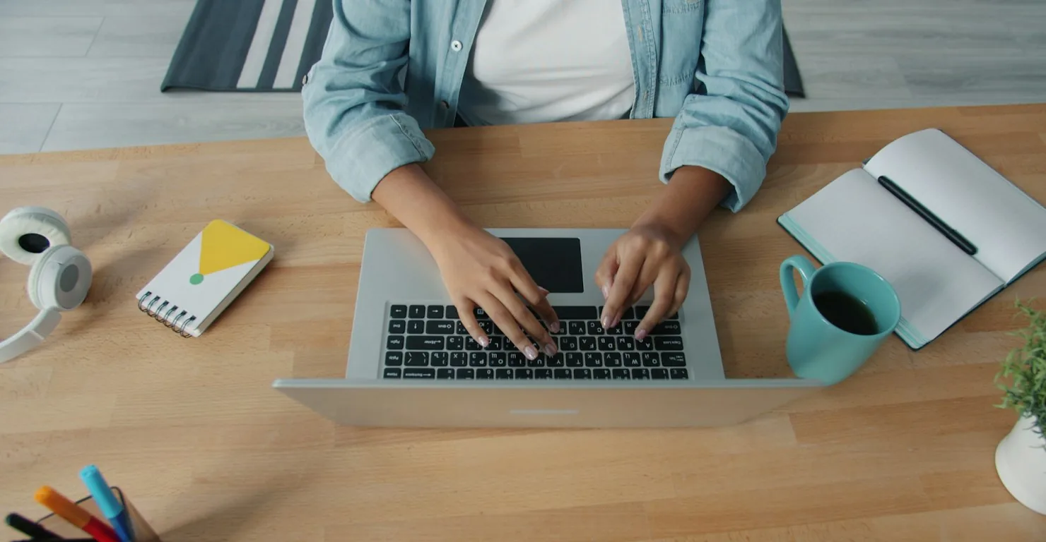 Mains d'une développeuse tapant sur un ordinateur portable posé sur un bureau en bois, avec carnet et tasse